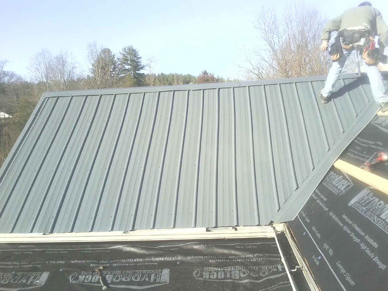 Crew installing dark green standing seam panels over synthetic underlayment in Olmstead