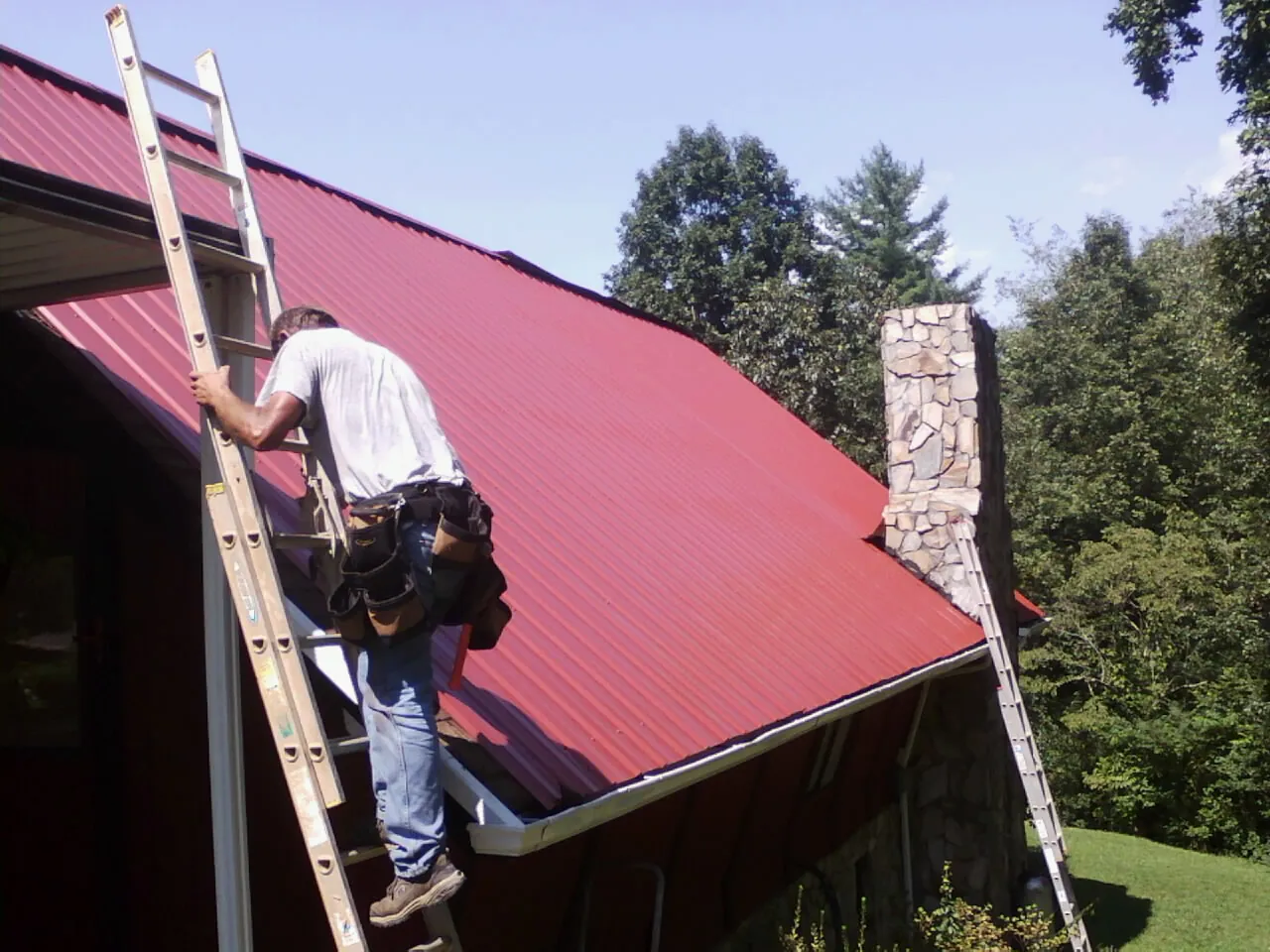 Roofer on a ladder beside a red metal roof and stone chimney — Soffit & Fascia Repair by Roofing Brothers in Olmstead